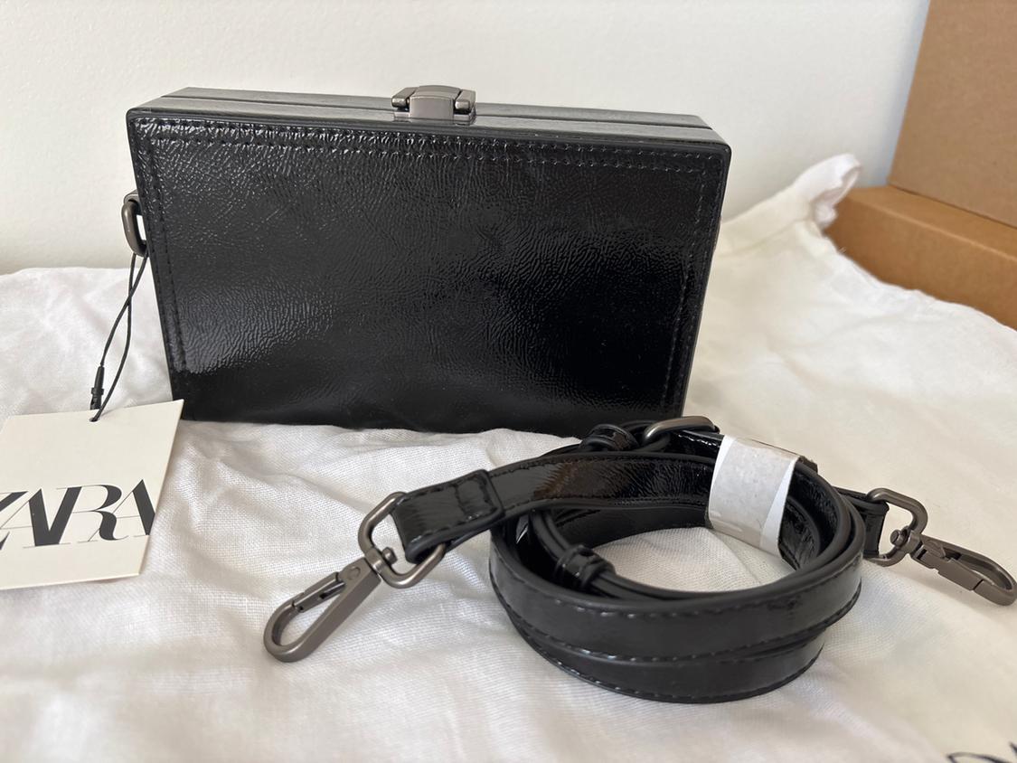 Rectangular black bag with detachable strap and Zara tag on white fabric. White wall background and cardboard box.