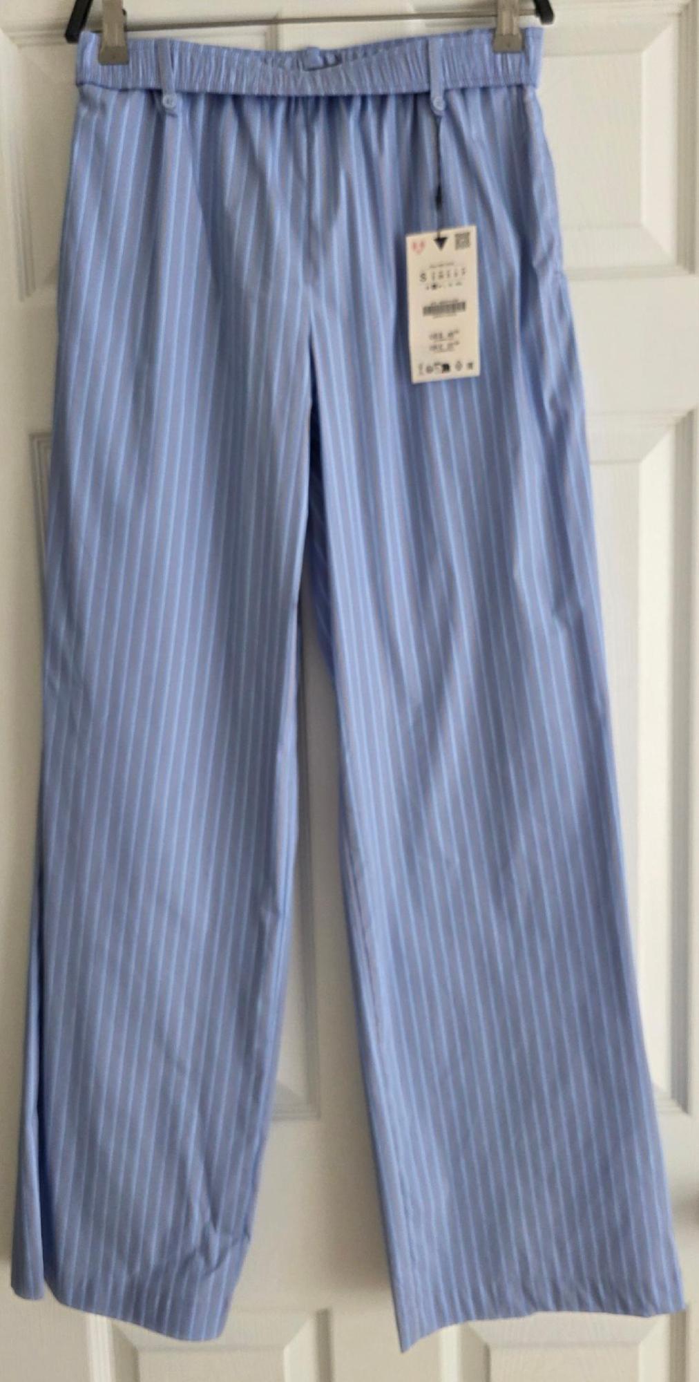 Light blue trousers with white vertical stripes, elasticated waistband and visible hanging tag, hanging in front of a white door.