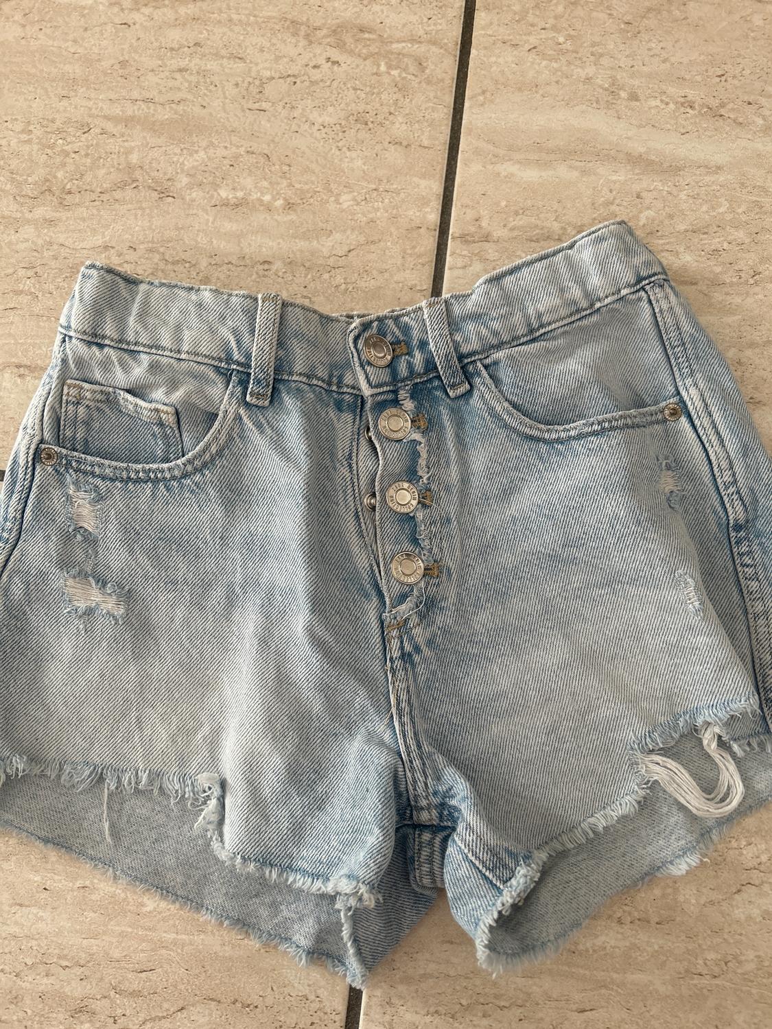 Light blue denim shorts with front buttons and distressed details, on a beige tiled background.