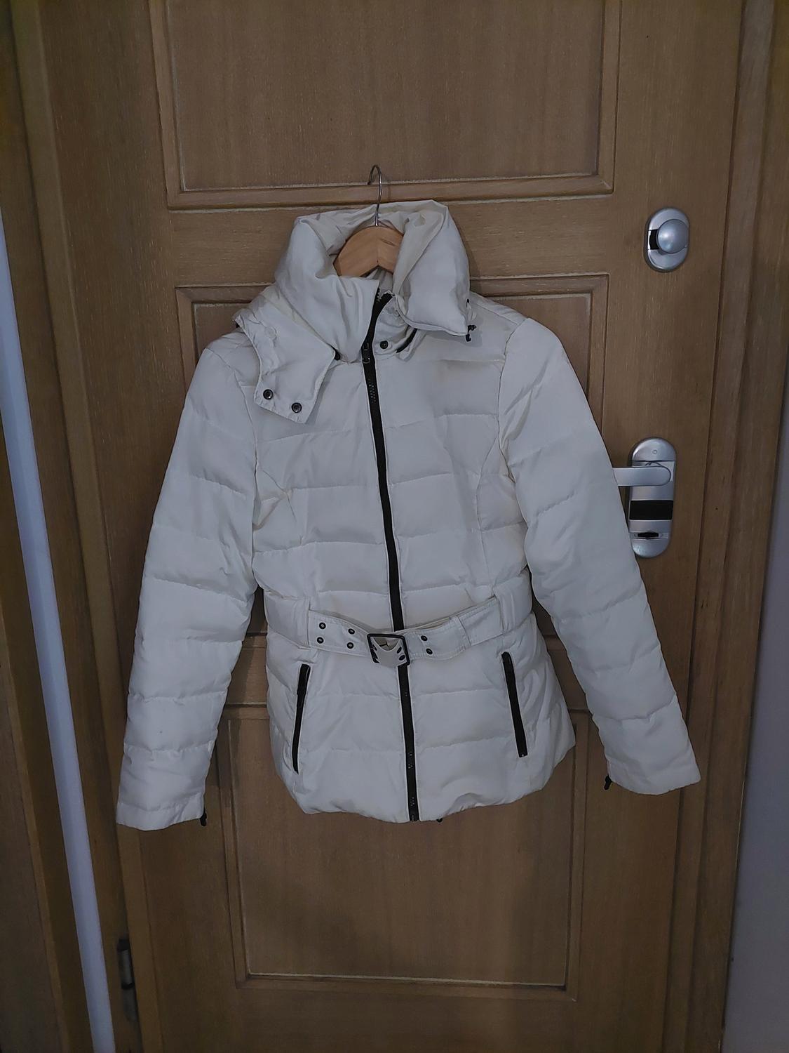 White padded jacket with belt and black zip, hanging on a wooden door.