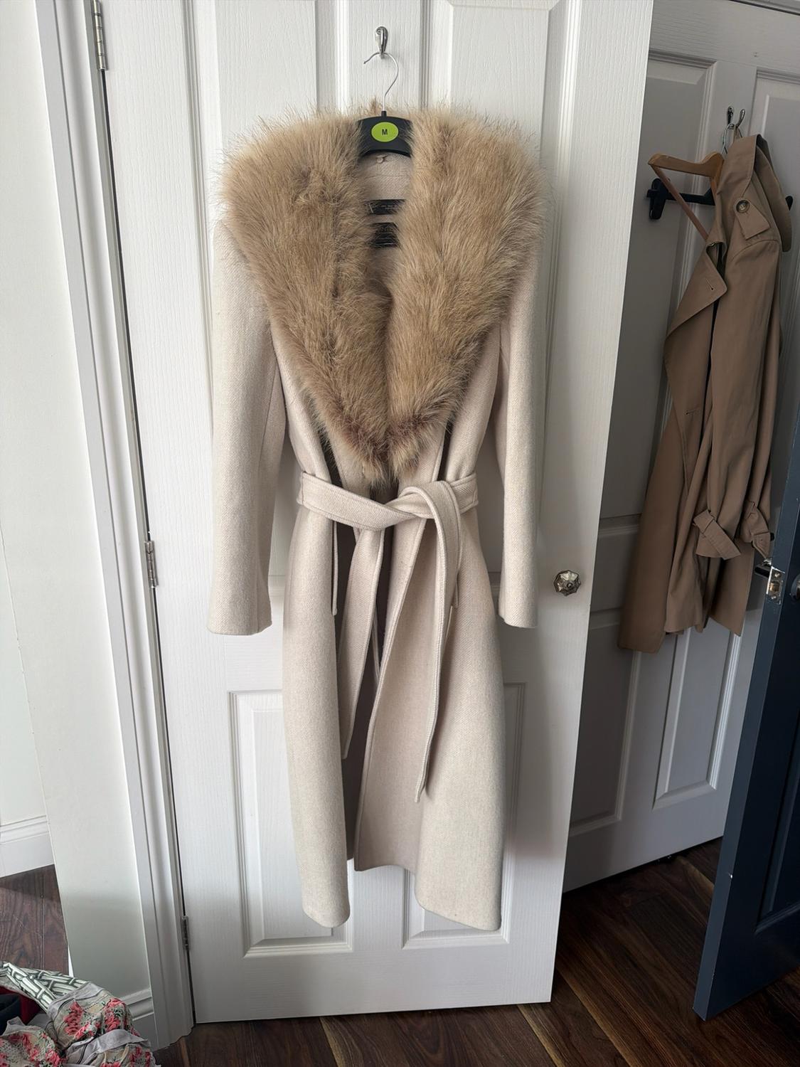 Long beige overcoat with a fur collar and a belt, hanging on a white door. The hanger has a green label with the letter M.