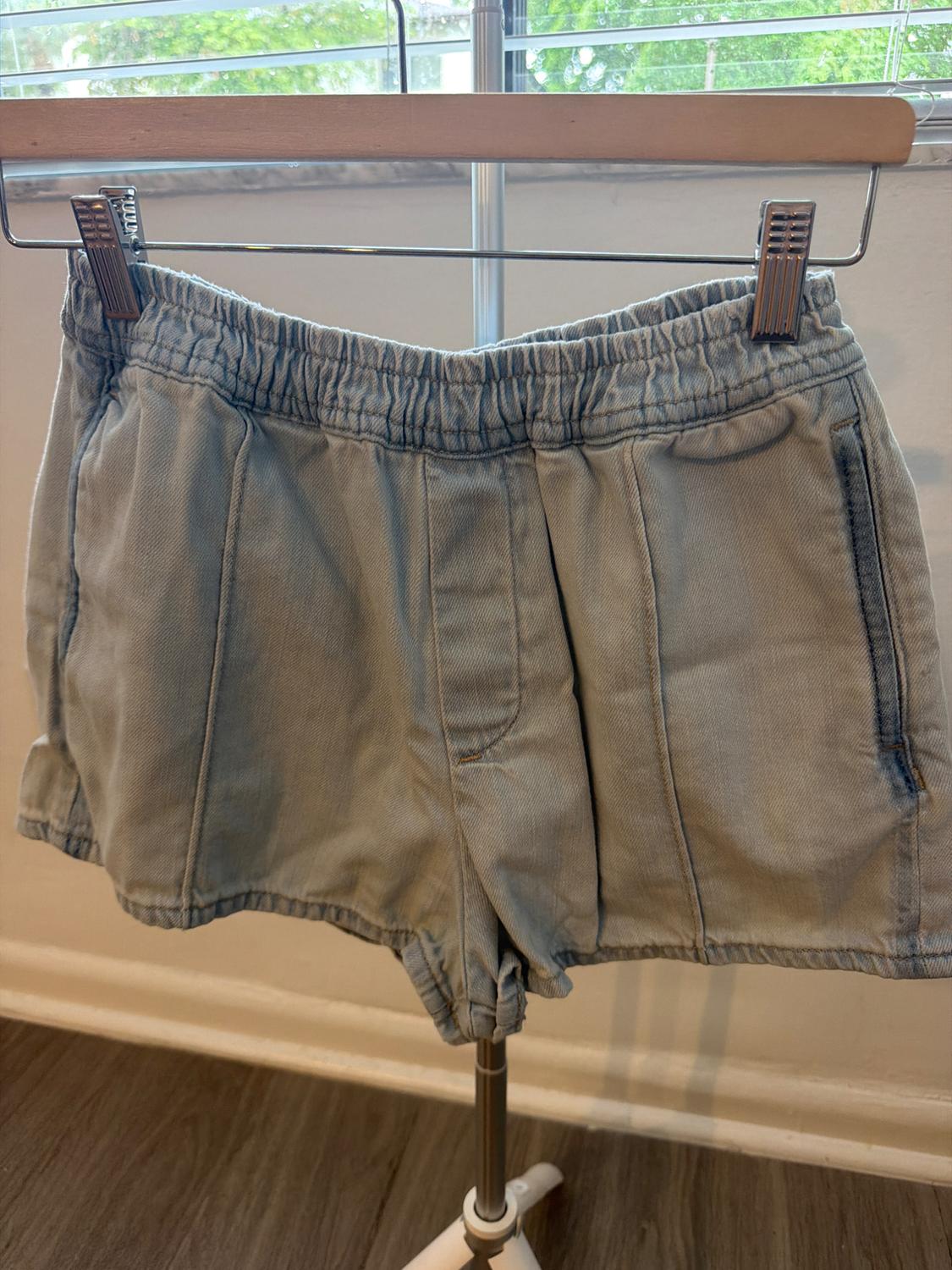 Light blue denim shorts with an elasticated waist, hanging on a clip hanger in front of a window with blinds.