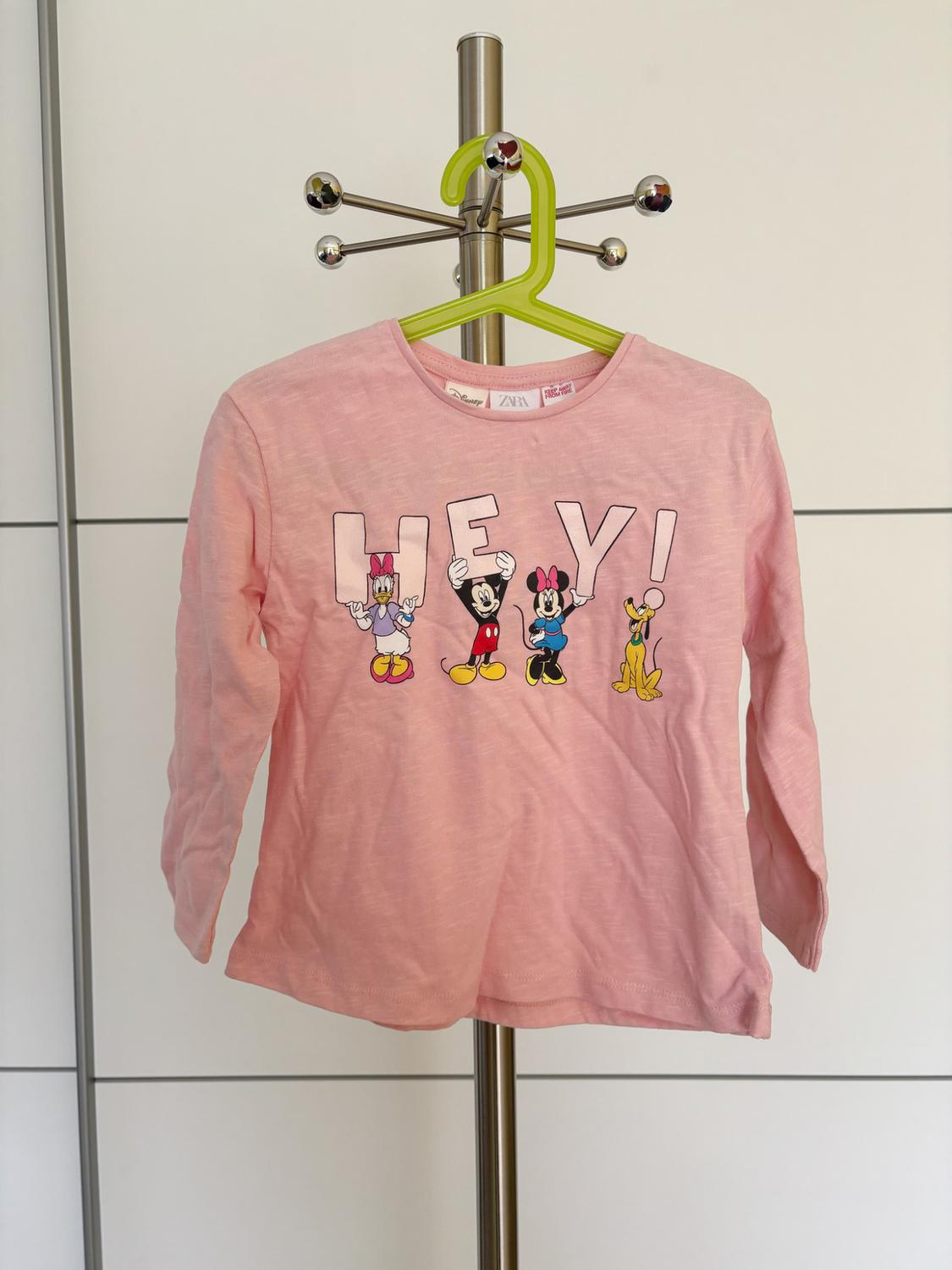 Ροζ μακρυμάνικο T-shirt κρεμασμένο σε μια πράσινη κρεμάστρα, με κινούμενα σχέδια και το κείμενο "HEY!" στο μπροστινό μέρος.