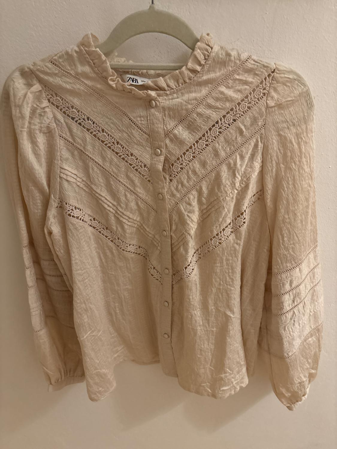 Beige long-sleeved blouse with lace and front buttons, hanging on a hanger. Visible label with text "ZARA".