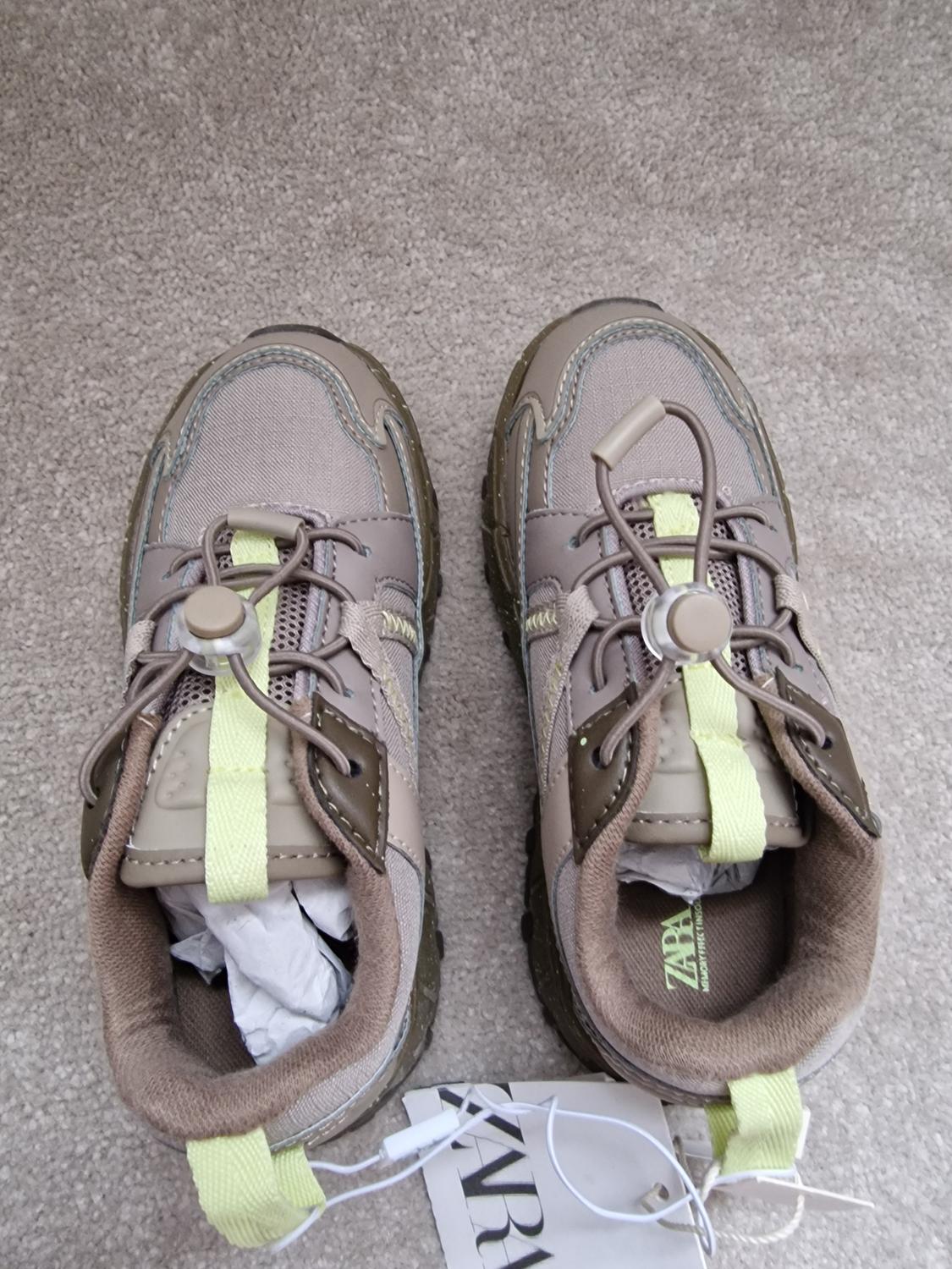 Grey trainers with green detailing and elasticated laces, on a beige carpet. Visible label with text "ZARA".