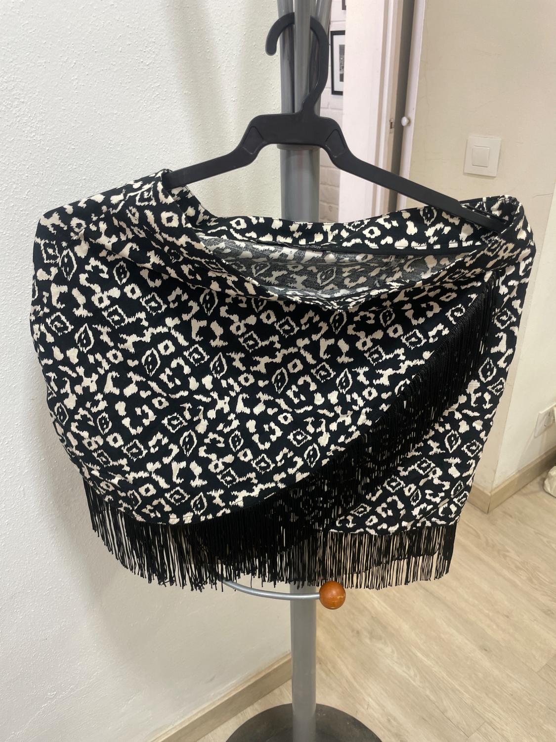 Black shawl with white print and tassels hanging on a hanger against a background of white wall and light wood floor.