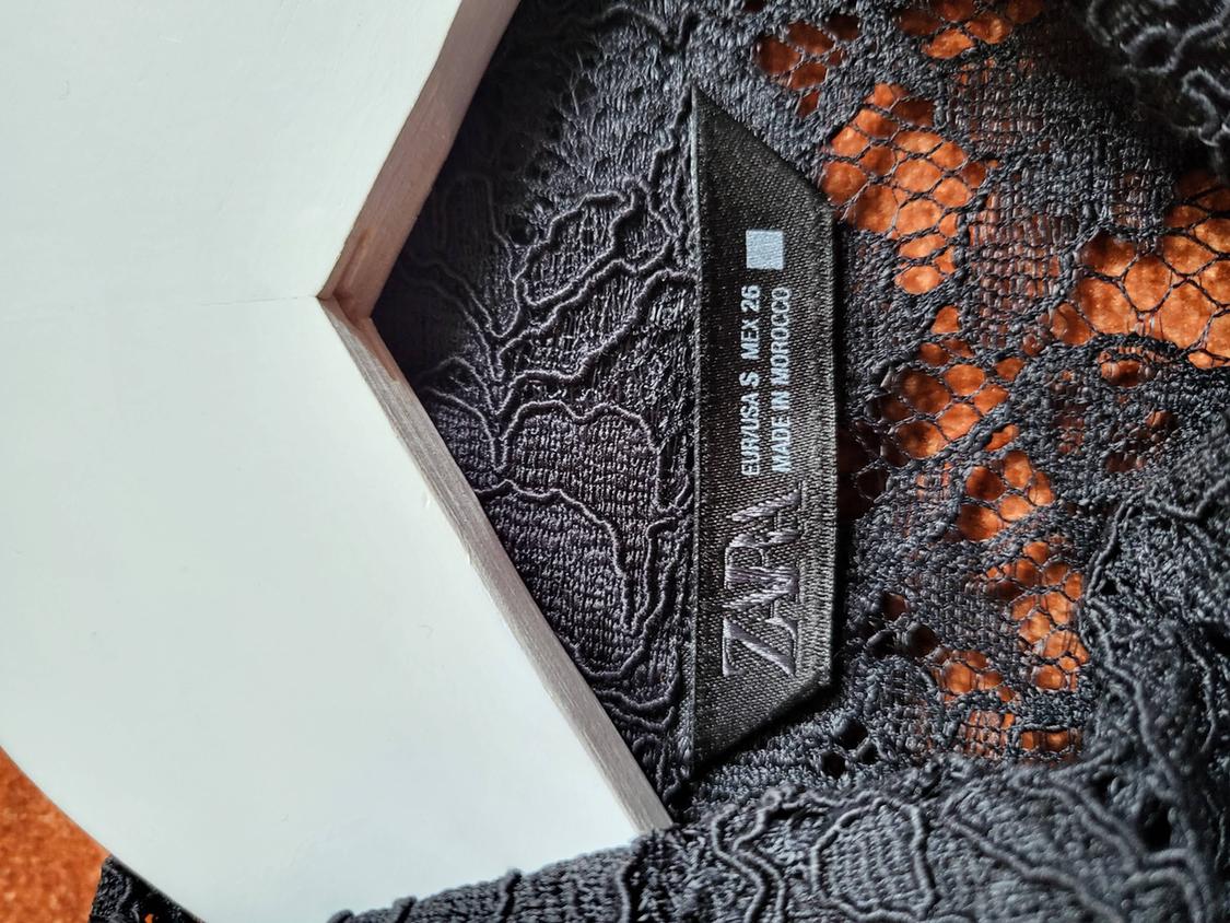 Black clothing label with lace, visible text: "ZARA EUR/USA: S MEX: 26 MADE IN MOROCCO", on a white and wooden background.