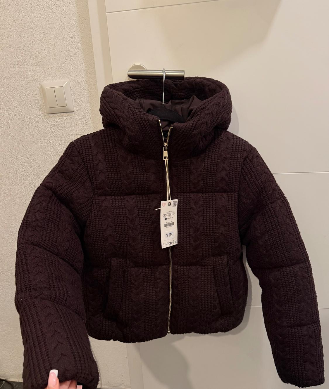 Dark brown padded jacket with a hood and zip, hanging on a hanger in front of a white door. Visible tag.