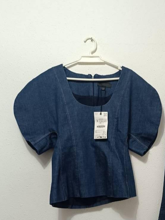 Blue denim top with a square neckline, pronounced seams and short puff sleeves, hanging on a hanger against a white wall.