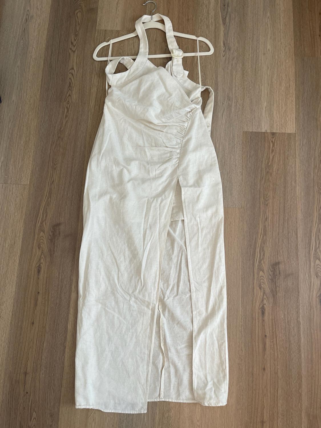 Beige color dress with a halter neckline, side slit, and ruched detail, hanging on a white hanger over a wooden floor.