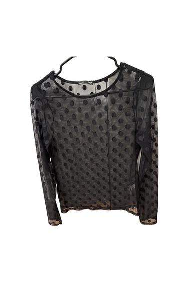 Sheer black blouse with large polka dots, long-sleeved, hanging on a hanger. Visible tag on the collar.