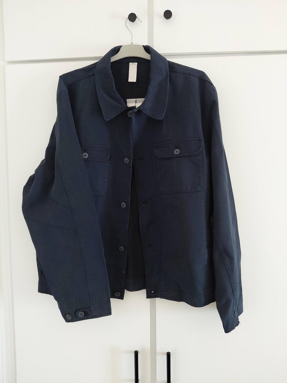 Navy-blue long-sleeved jacket with a collar, buttons and chest pockets, hanging on a hanger above a white wardrobe.