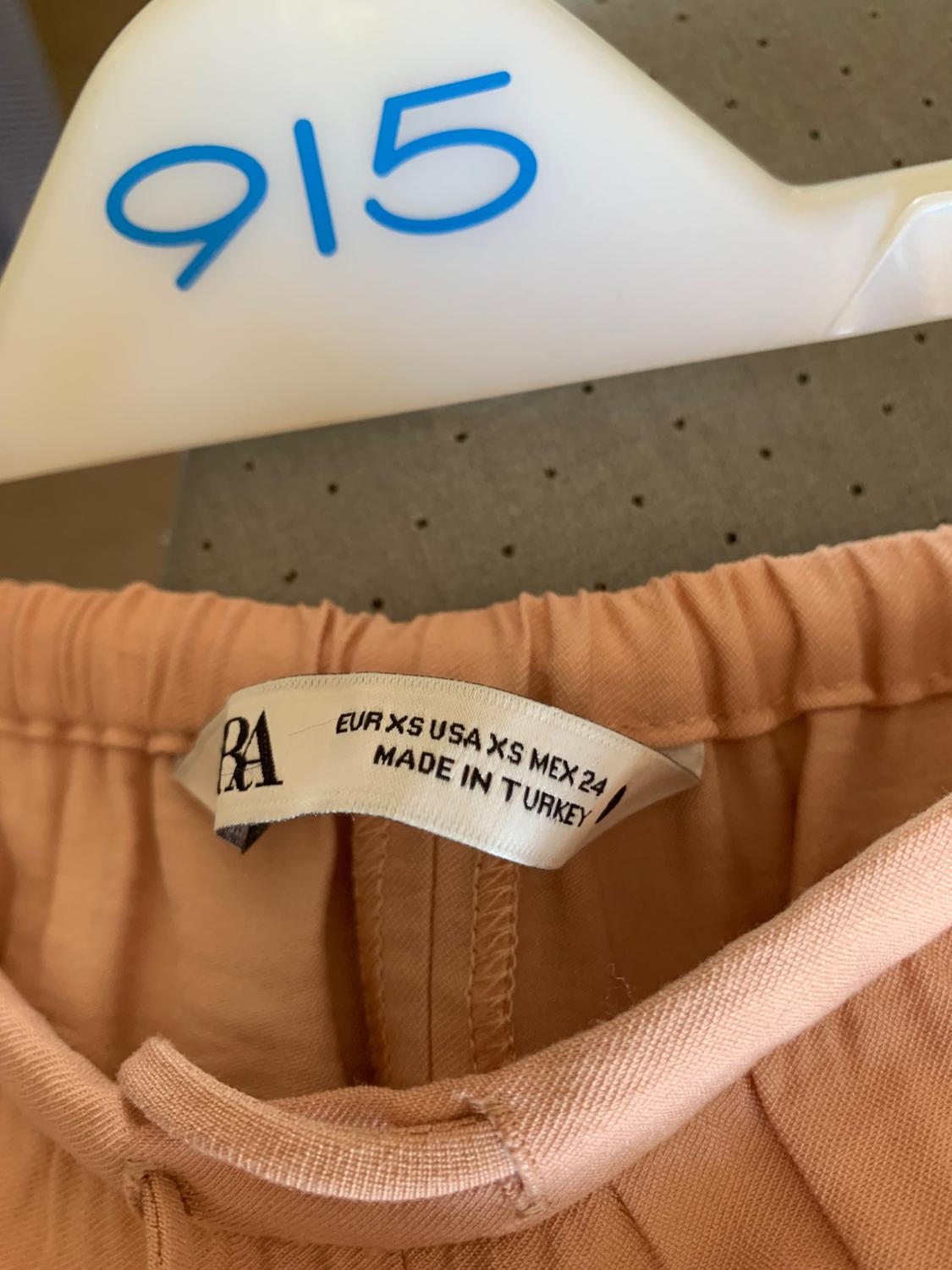 Beige coloured trouser label on a white hanger with the blue number "915". Text on label: "EUR XS USA XS MEX 24 MADE IN TURKEY".