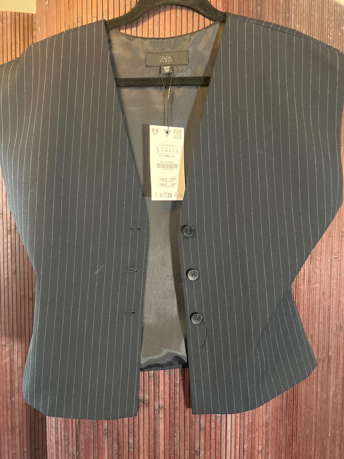 Black vest with vertical stripes hanging on a hanger, with a visible tag showing the price and size. Wooden background.
