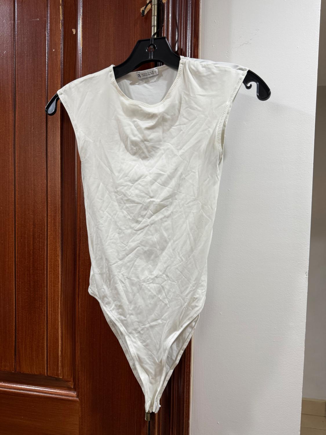 Sleeveless white bodysuit hanging on a black hanger over a wooden door. Wrinkled fabric and crew neck.