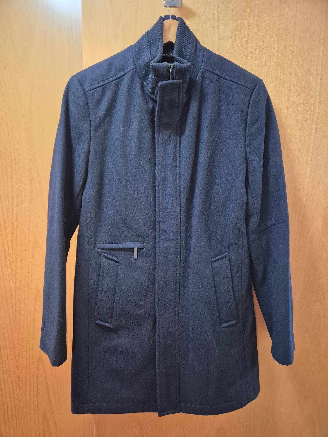 Dark blue coat hanging on a wooden hanger over a light wooden door. Simple design with front pockets.