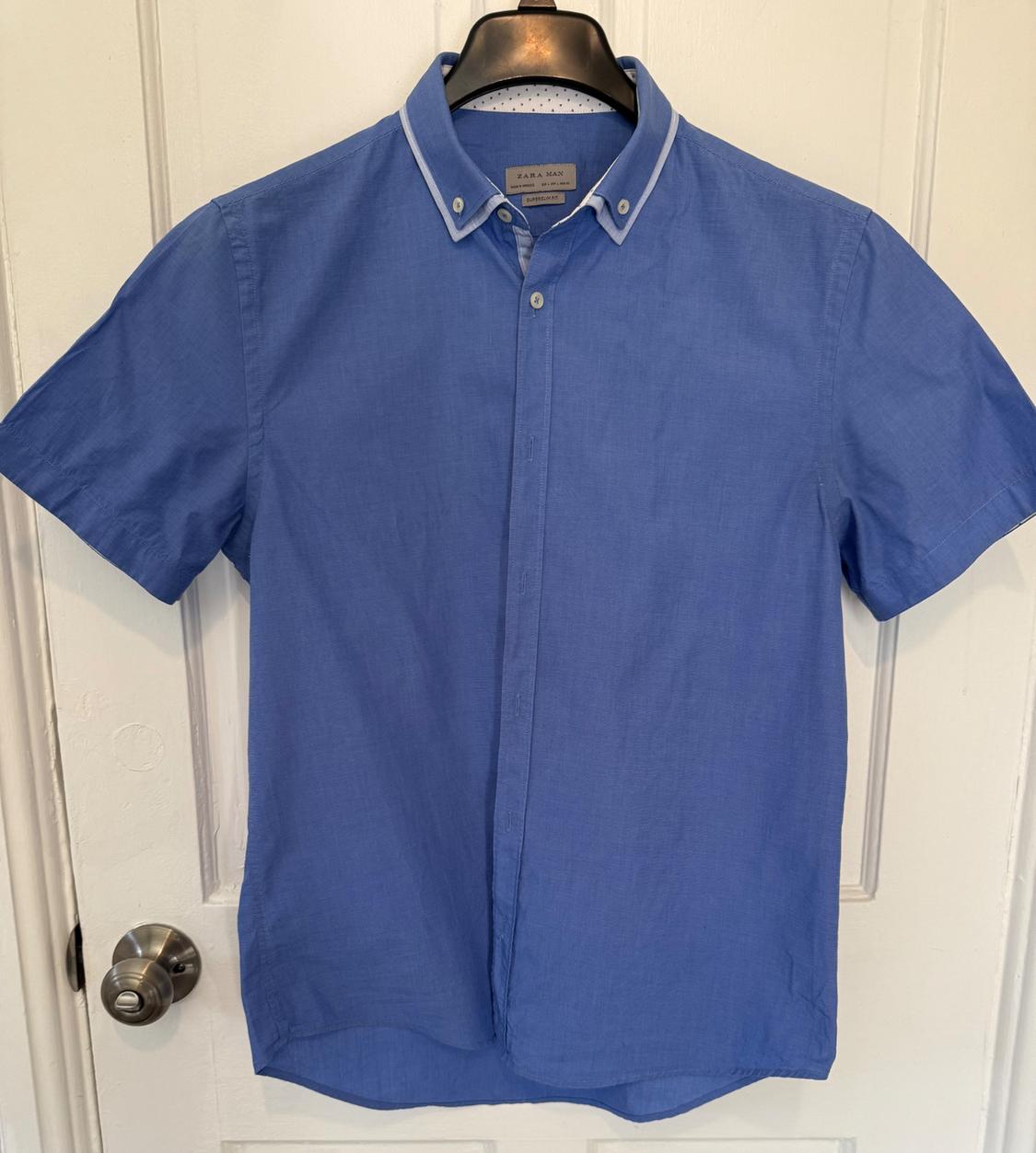 Blue short-sleeved shirt hanging on a black hanger over a white door. Visible label: "Zara Man".
