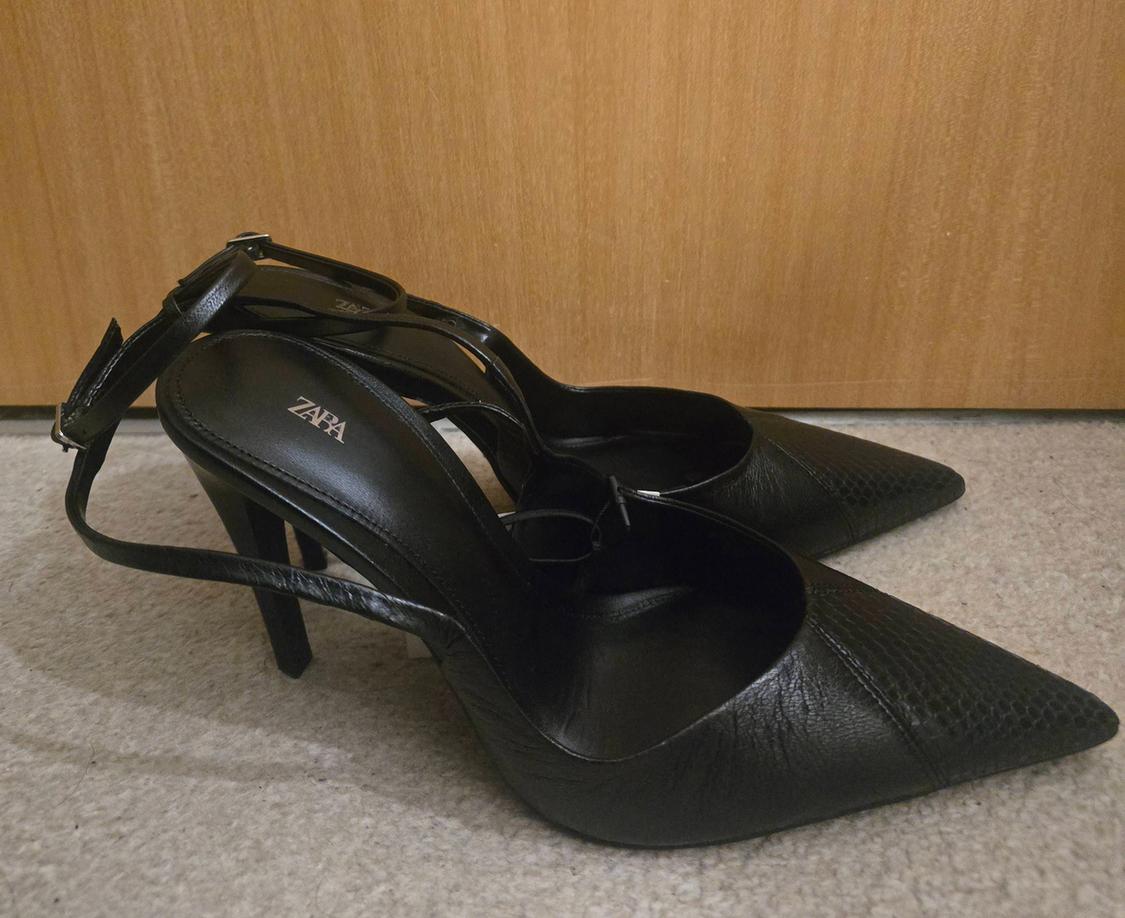 Black high-heeled shoes with a pointed toe and adjustable strap, Zara brand, on a beige carpet and light wood background.