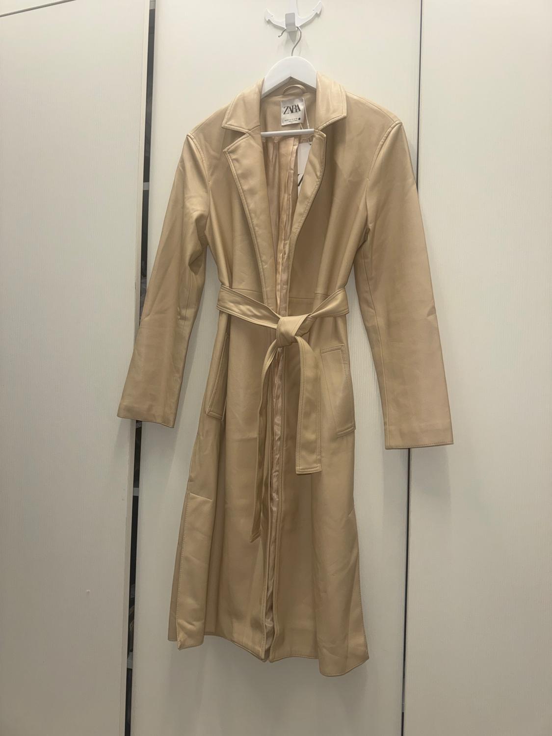 Long beige coat with belt, hanging on a white hanger. Visible label with the text "ZARA". White wall background.