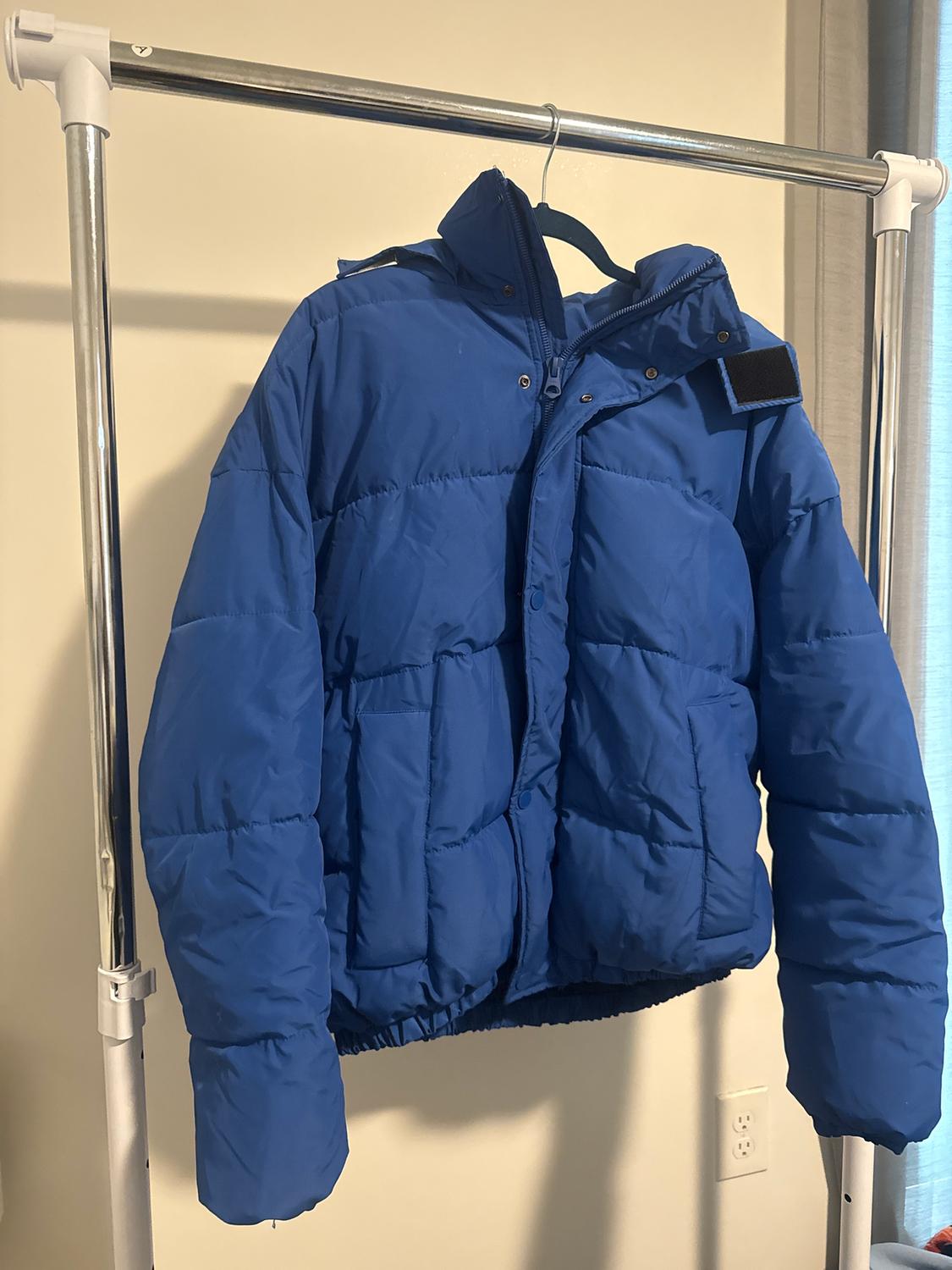 Blue padded jacket hanging on a metal coat rack. Beige wall background and light blue curtain on the right.