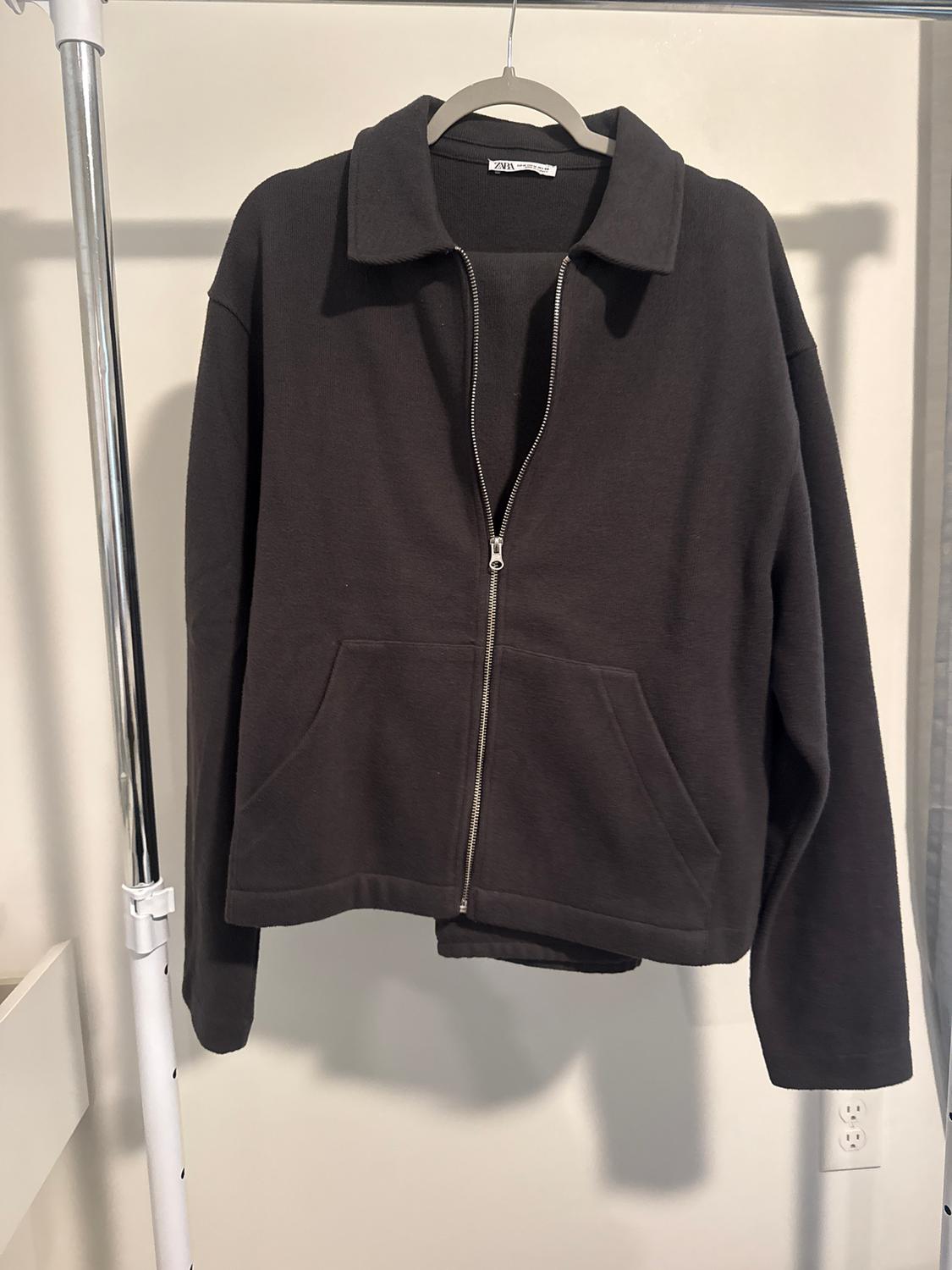 Black jacket with zip and front pockets, hanging on a hanger against a white background. Label visible on collar.
