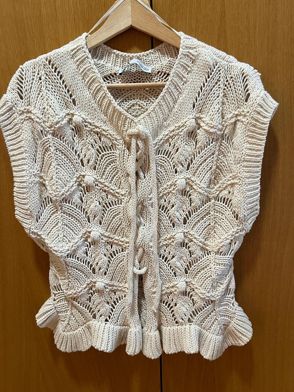 Ecru openwork knit waistcoat with raised motifs, front tie fastening and ruffled hem, hanging on a wooden hanger.