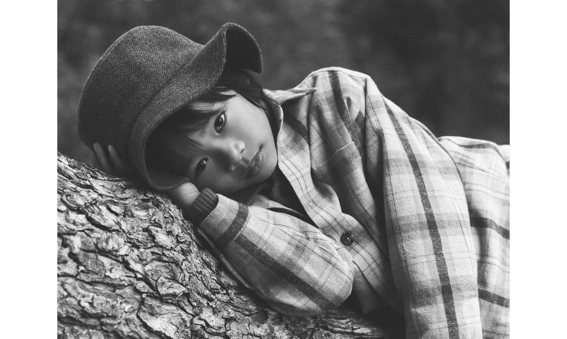 Black and white image of a boy lying on a log, wearing a checked shirt and hat, looking at the camera with a calm expression.