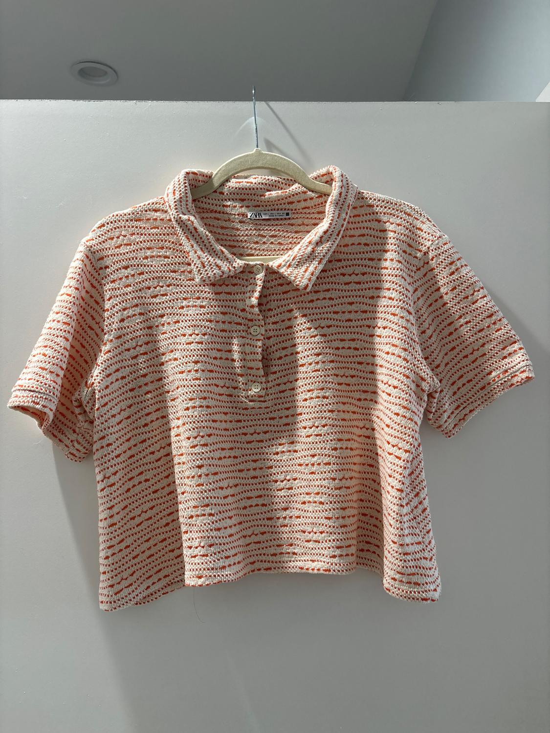 Knitted shirt with collar and buttons, in beige and orange tones, hanging on a hanger against an illuminated white background.