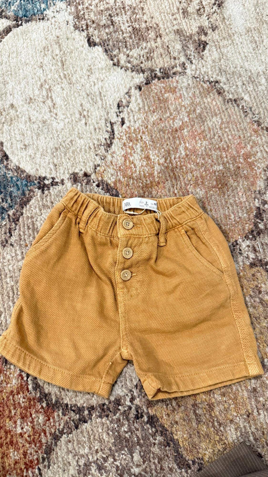 Light brown shorts with wooden buttons on textured carpet in beige and brown tones. Label visible at the waist.