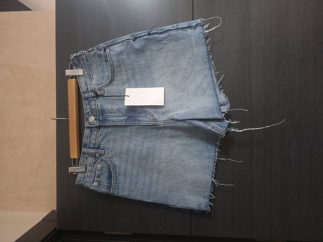 Ανοιχτόχρωμο μπλε τζιν σορτς με ξεφτισμένο τελείωμα σε κρεμάστρα πάνω σε σκούρο ξύλινο φόντο. Ετικέτα με κείμενο ZARA.