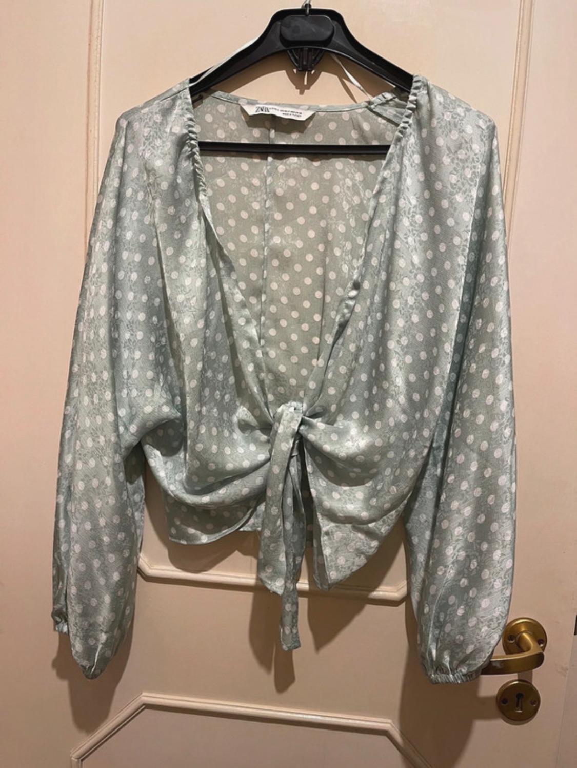 White polka dot blouse on a light green background, with a bow at the waist, hanging on a black hanger over a beige door.