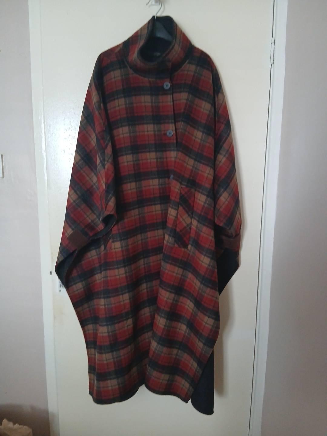 Red, black and brown checked cape hanging on a white door. It features a high neck, front buttons and a pocket.