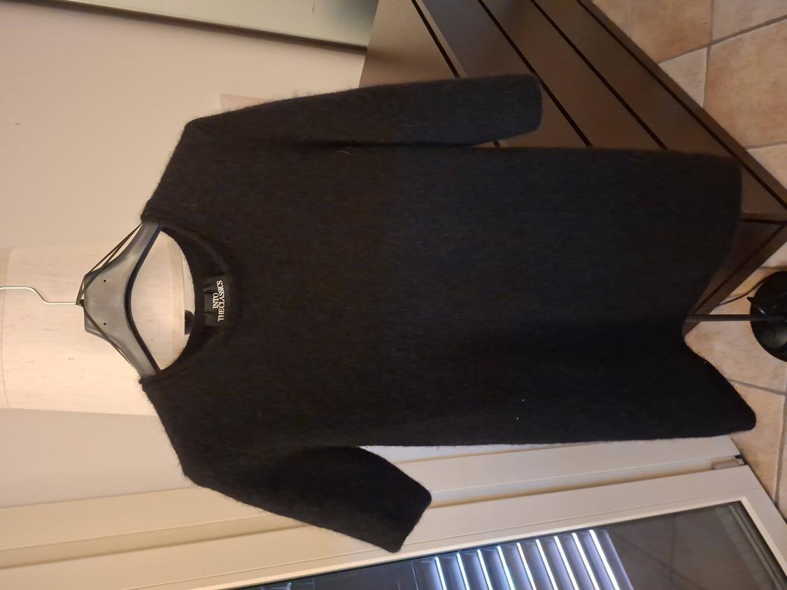 Black three-quarter sleeve jumper hanging on a hanger against a background of furniture and tiled flooring. Visible tag: "INTO THE CHAOS".