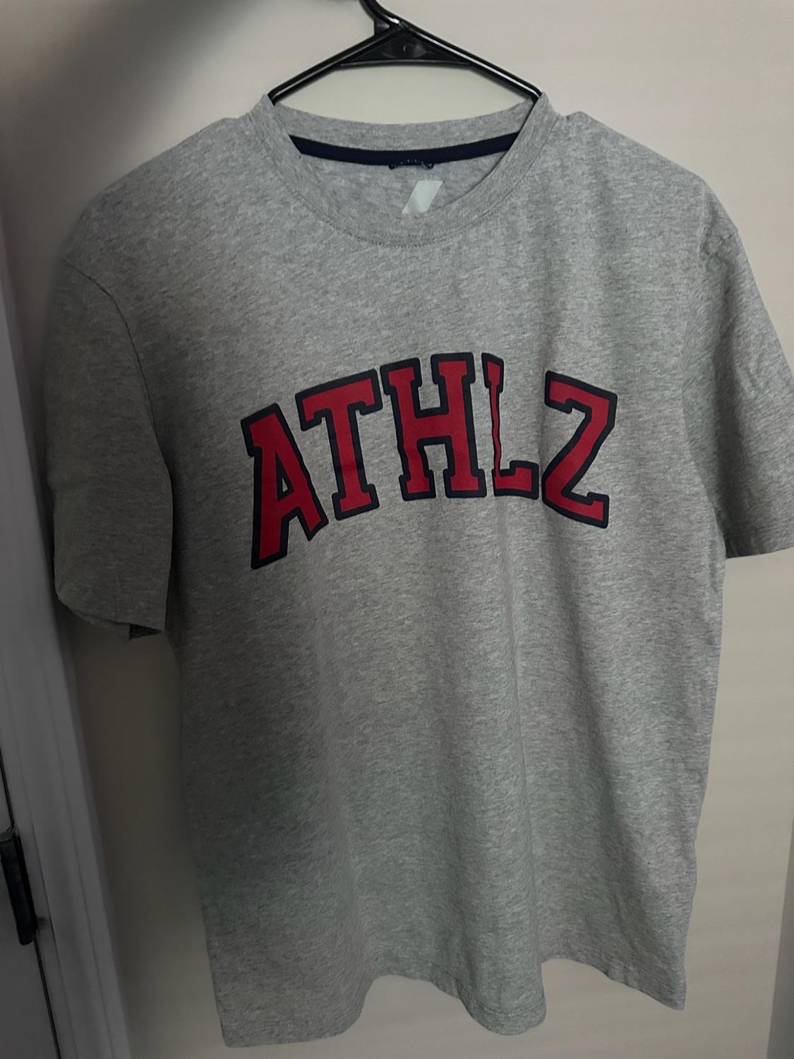 Grey short-sleeved T-shirt hanging on a hanger. The text “ATHLZ” is printed on the chest in arched red letters.