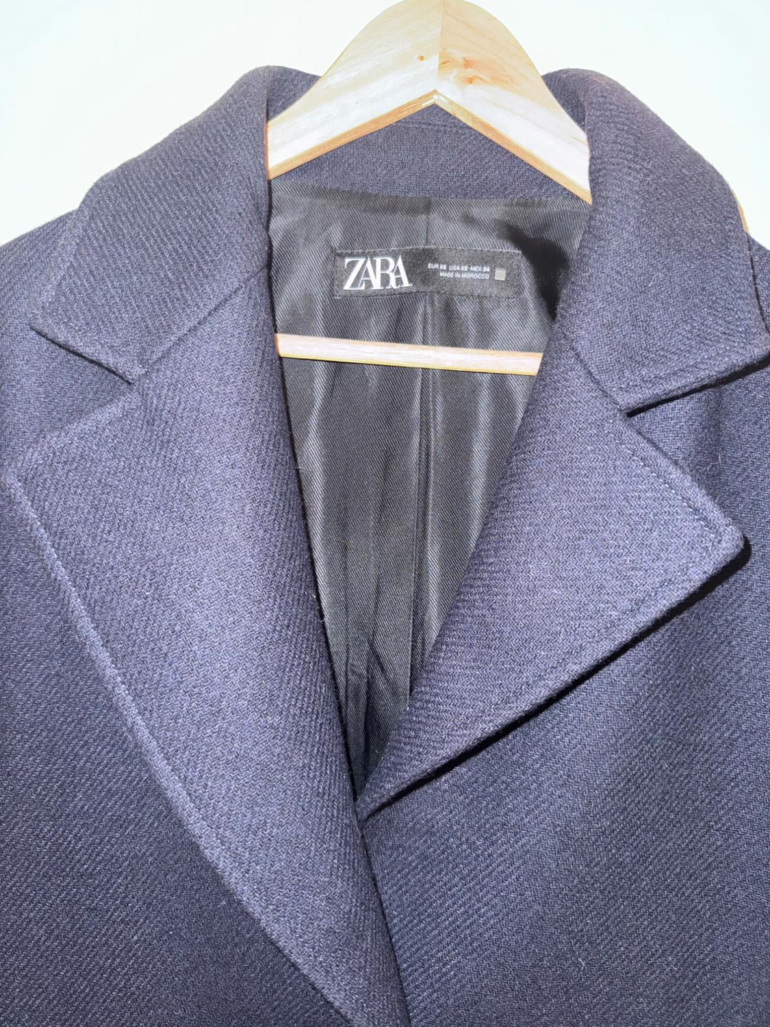Dark coat hanging on a wooden hanger, with a visible label that says "ZARA". Textured fabric and black inner lining.