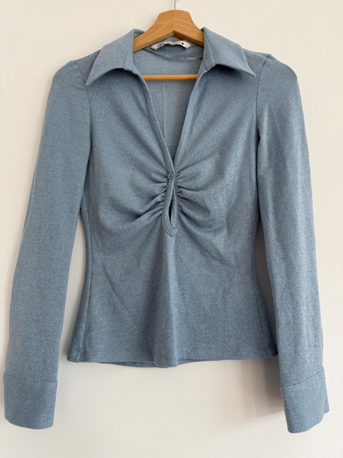 Light blue shirt with a wide neck and central ruching, long-sleeved, hanging on a wooden hanger against a white background.