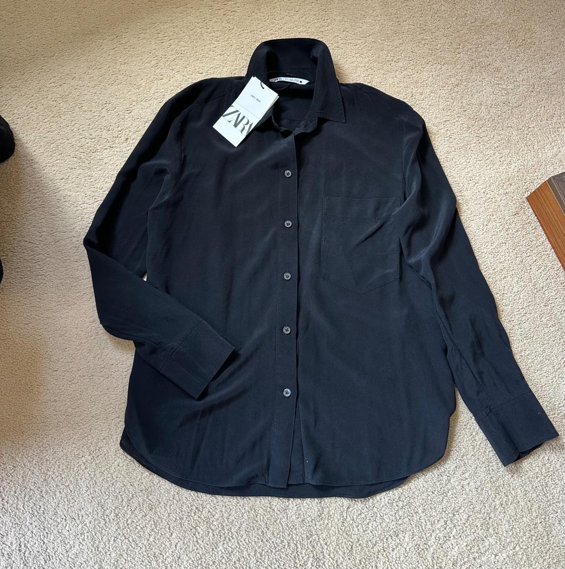 Black long-sleeved shirt with buttons and a pocket, laid out on a beige rug. It has a white label with the text ZARA.