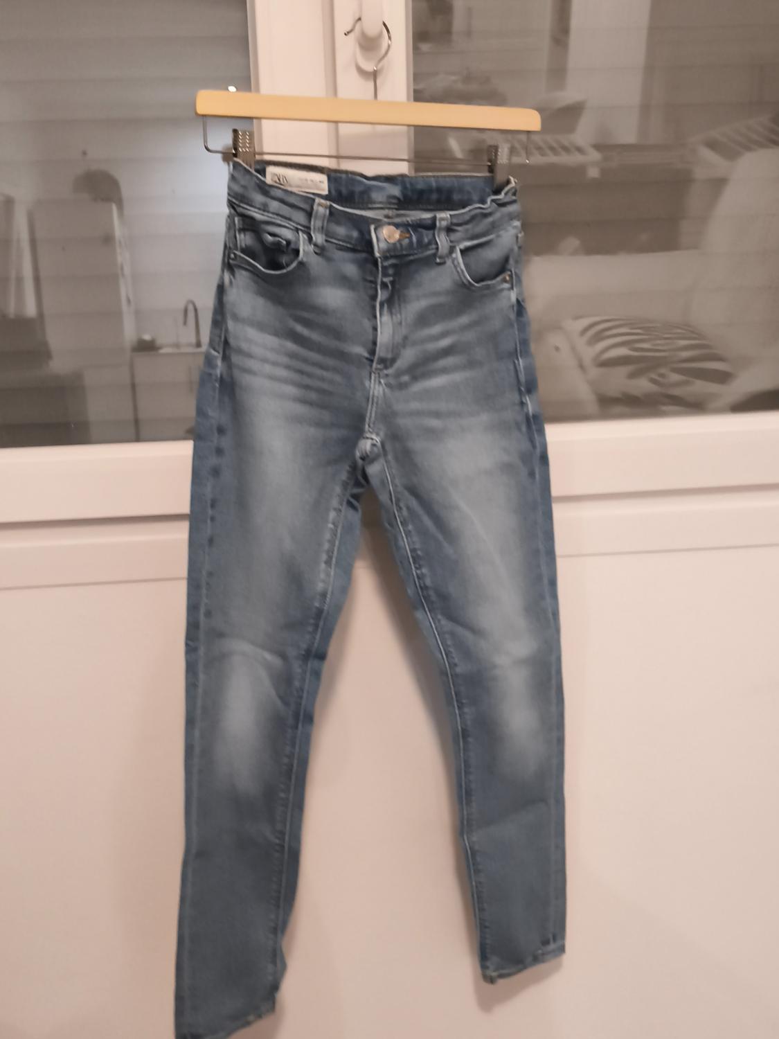 Light blue jeans hanging on a hanger in front of a window. Blurred indoor background with furniture visible.