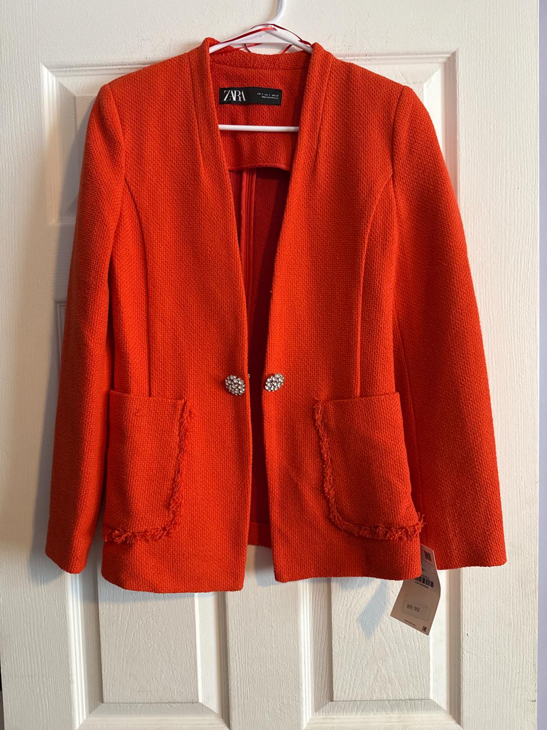 Orange textured fabric jacket with front pockets and decorative buttons, hanging on a white door. Visible label.