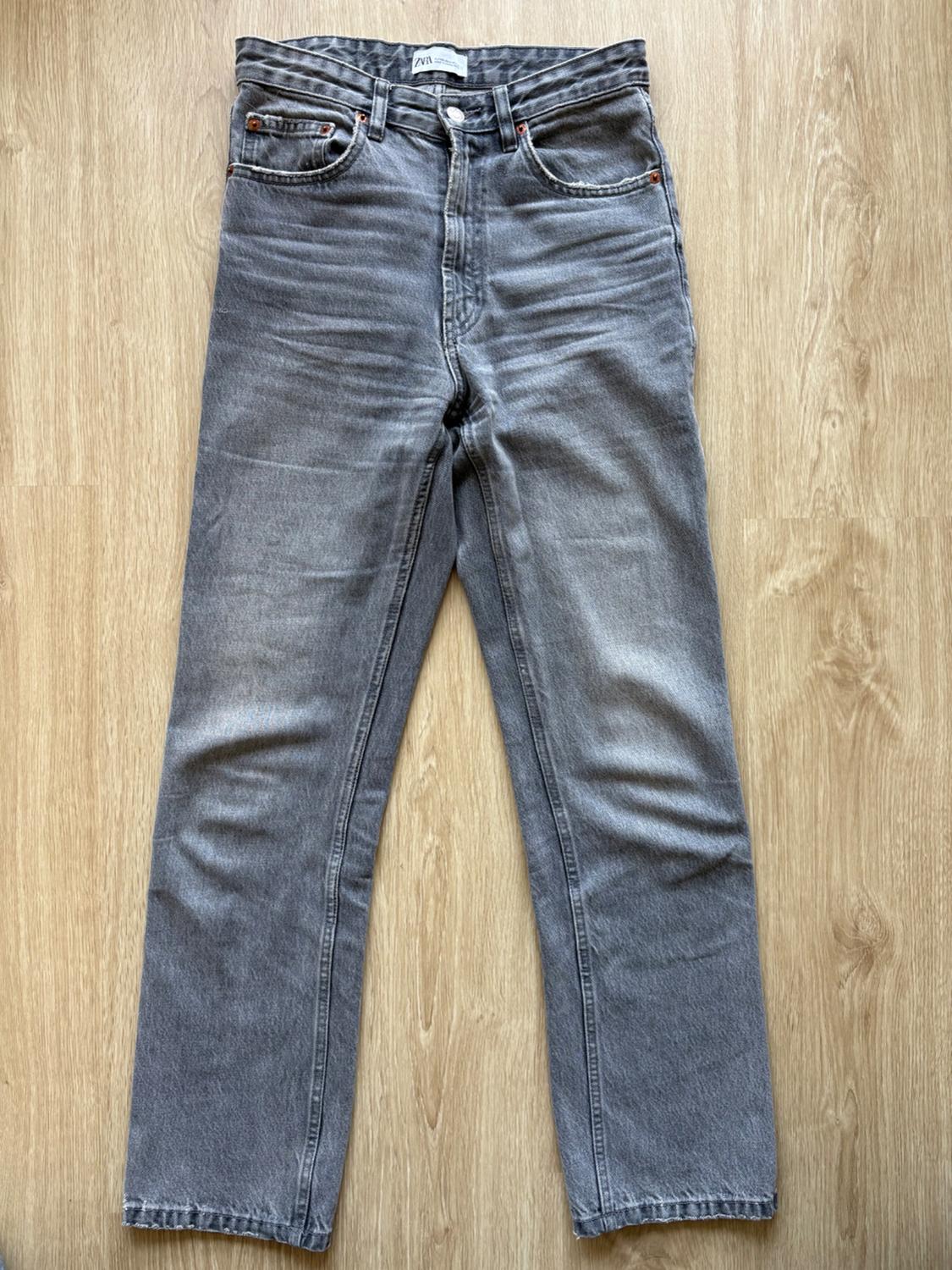 Grey straight-cut jeans on a light wooden floor. Visible label on the waistband with the text "ZARA".