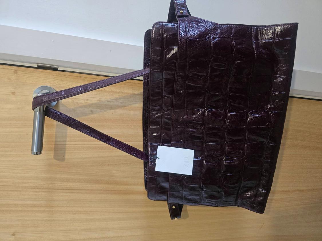 Glossy burgundy handbag with a crocodile texture hanging on the metal knob of a light wood door.