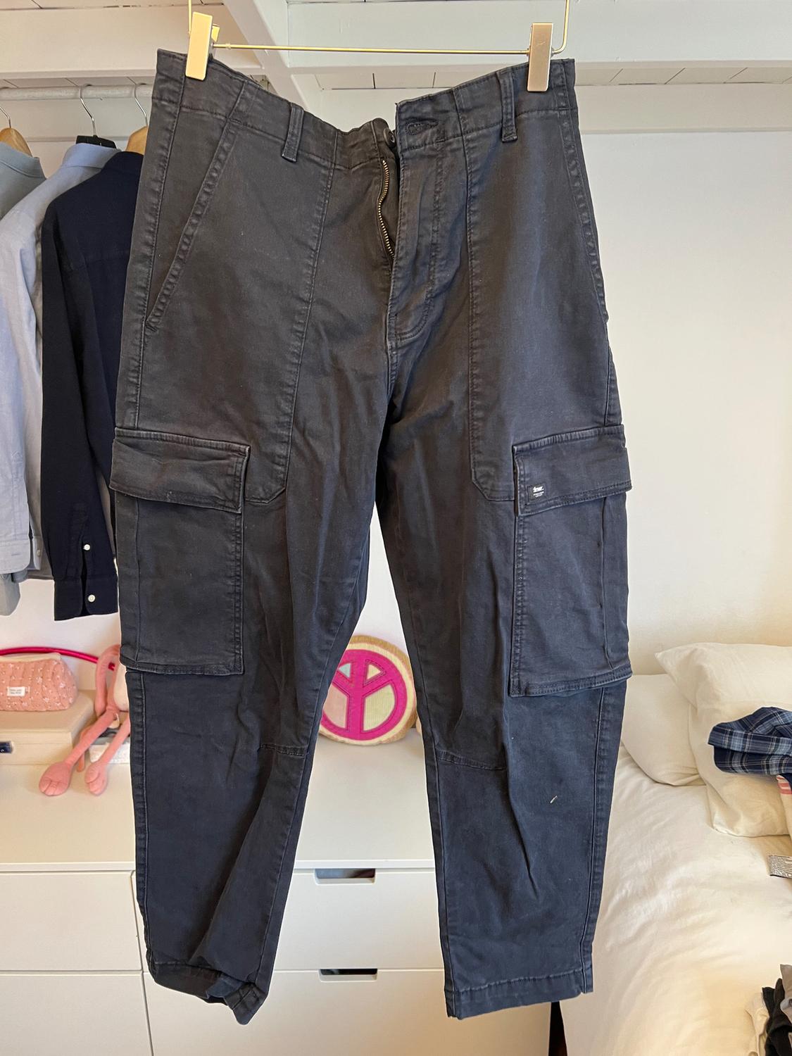 Black cargo trousers hanging on a hanger, with side pockets. Background with clothes hanging and a bed with cushions.