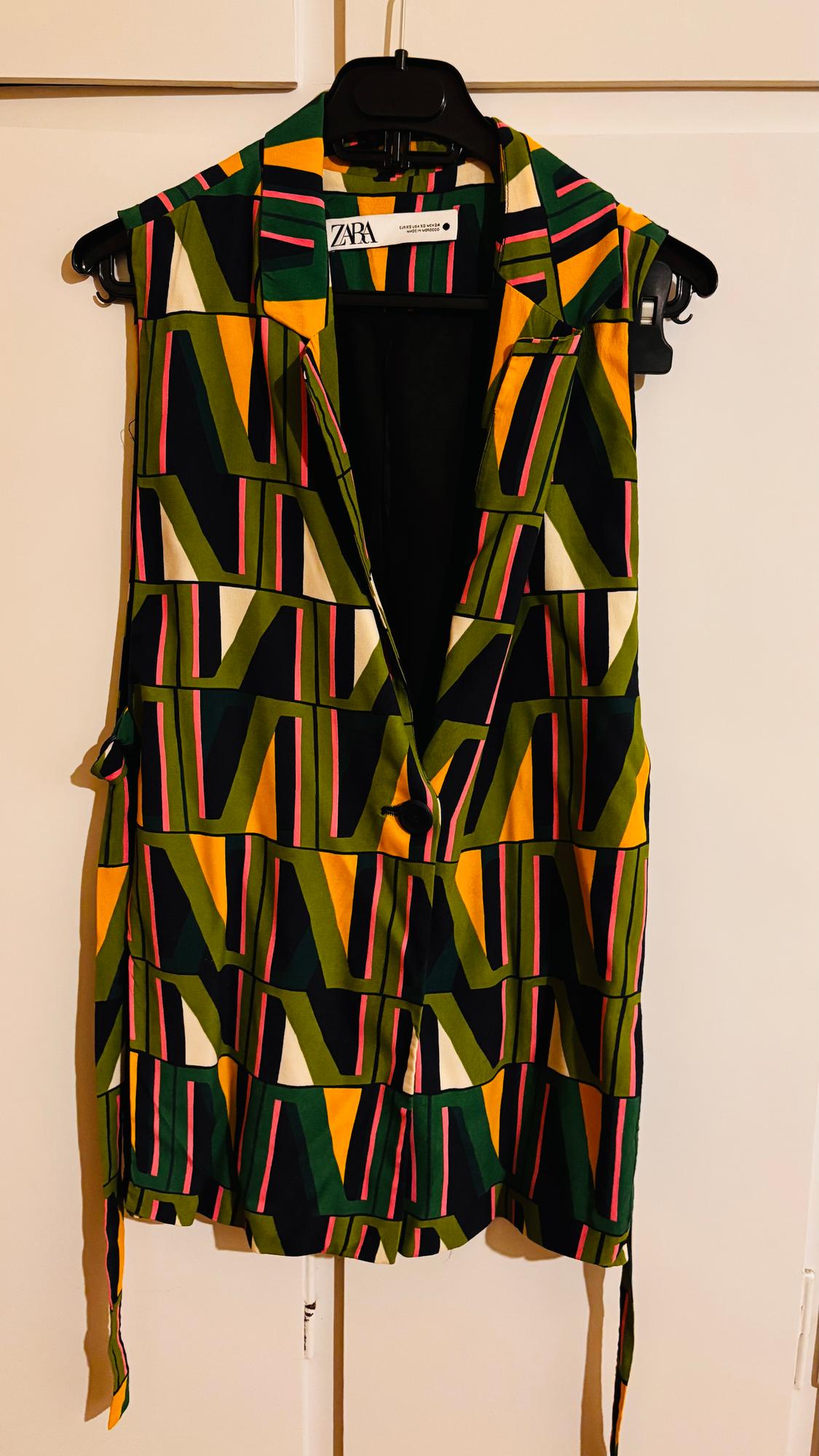 Sleeveless waistcoat with geometric print in green, yellow, and pink, hanging on a black hanger. Visible label: ZARA.