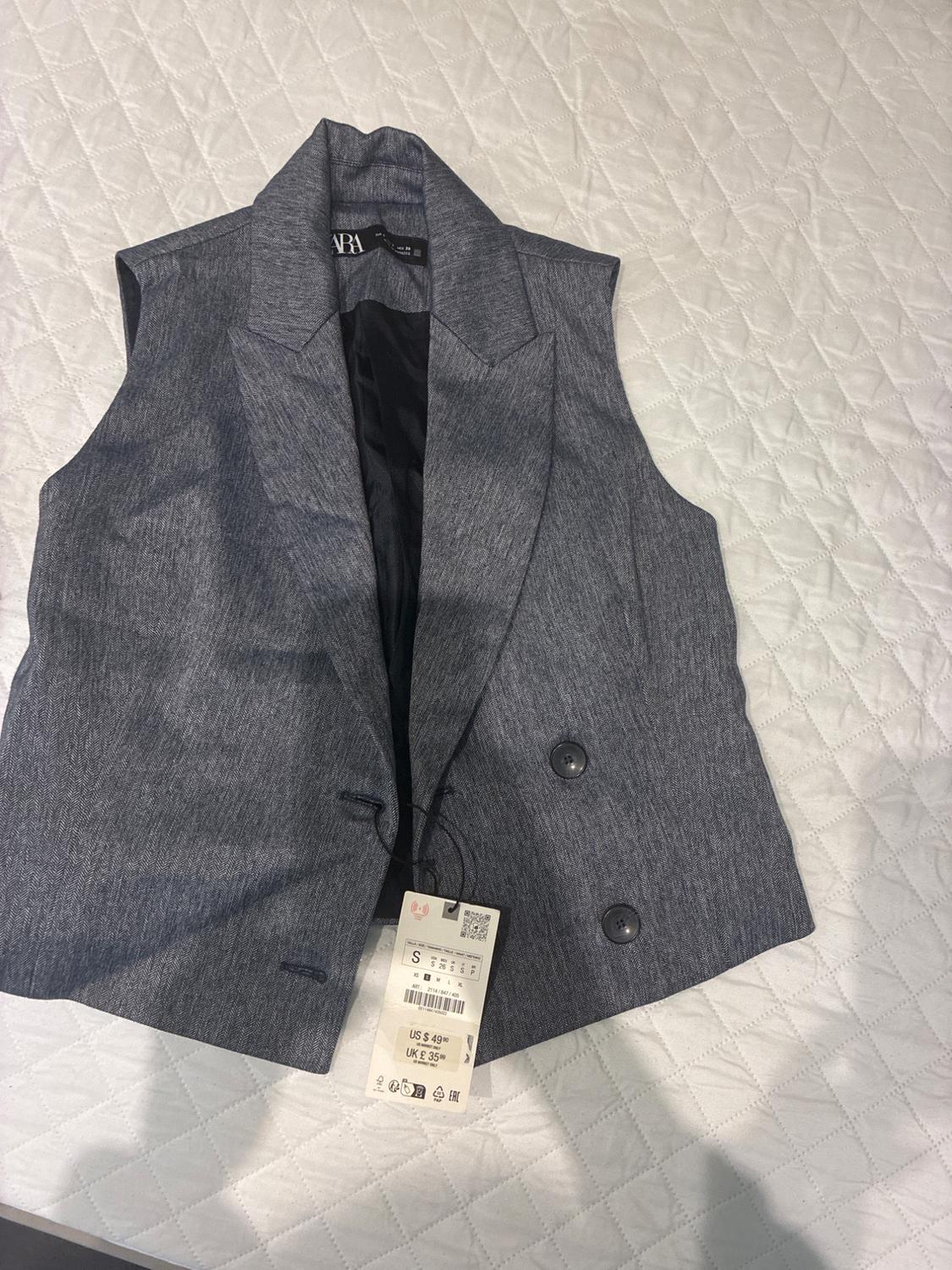 Sleeveless grey waistcoat with large lapels and two buttons, over a quilted white bedspread. Visible tag with price and size.