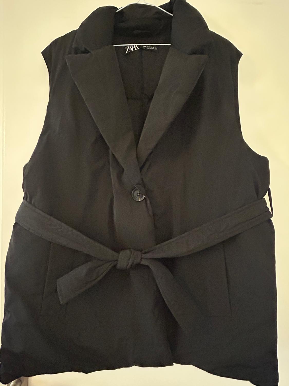 Black sleeveless waistcoat with a knotted belt at the front and wide lapels, hanging on a white hanger.