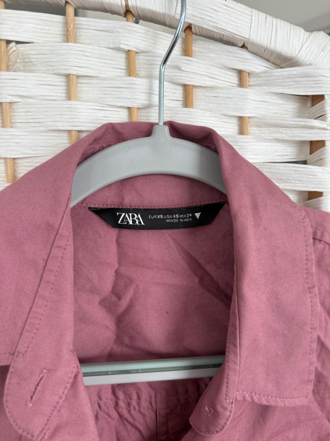 Pink shirt on a grey hanger with a Zara tag, hanging in front of a white woven basket background.