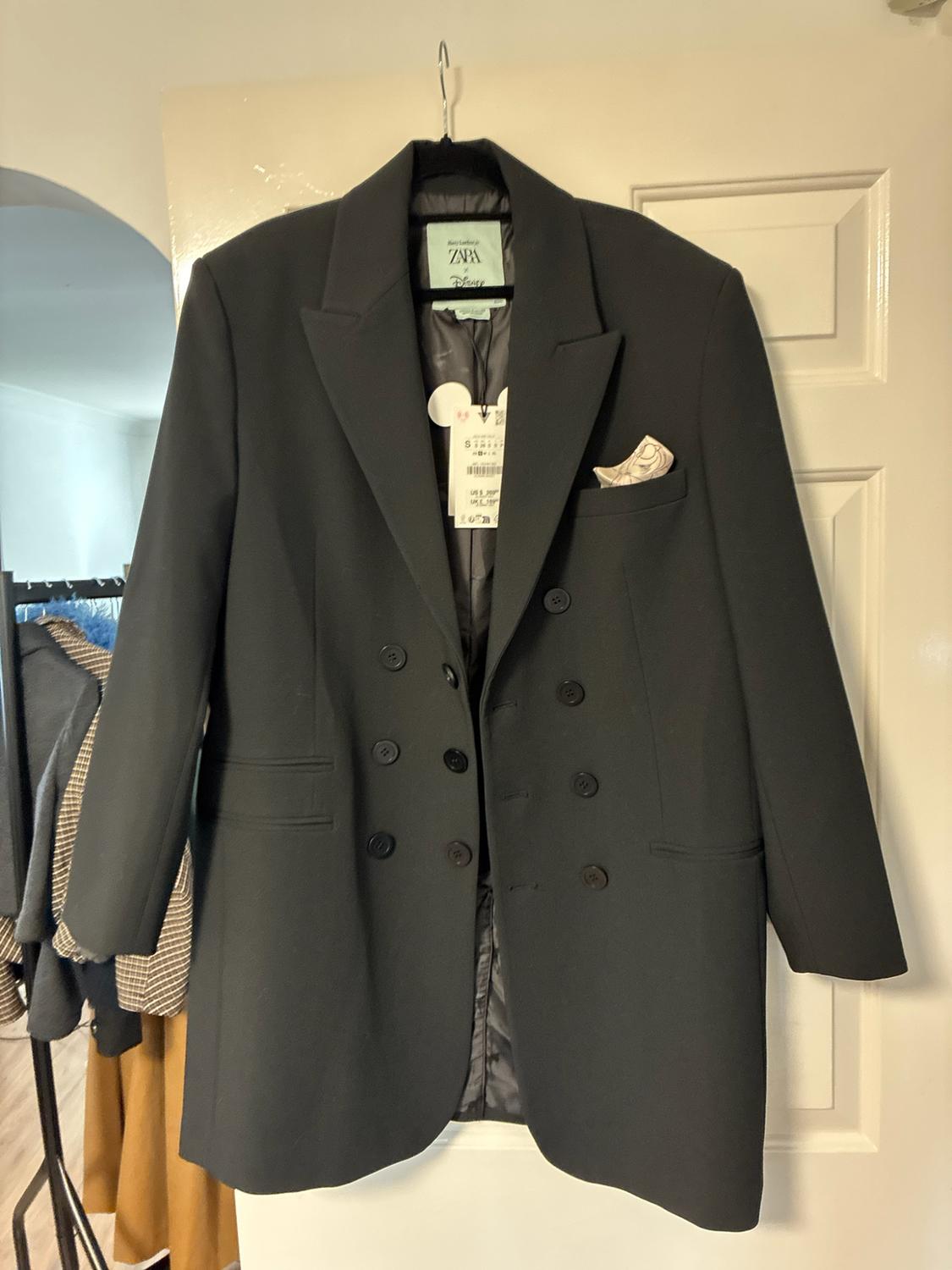 Black double-breasted blazer hanging on a white door. Label with the text ZARA and Disney. Handkerchief in the pocket.