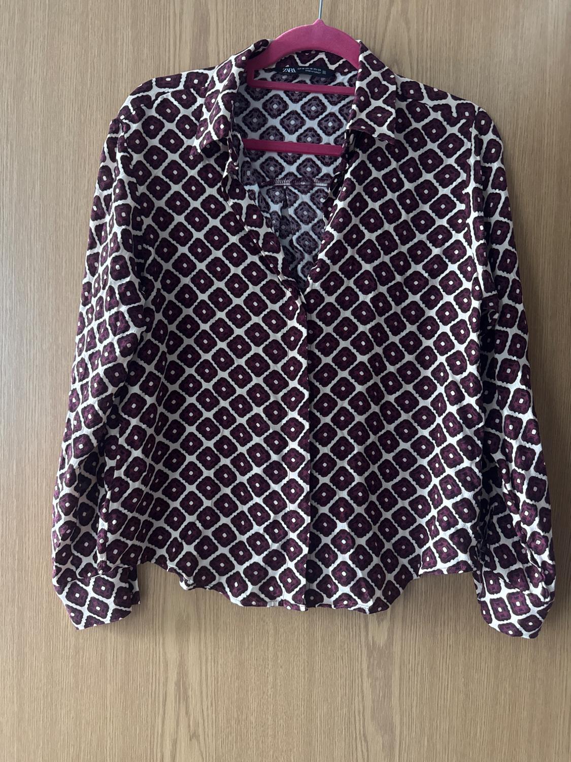 Maroon and white geometric print long-sleeve button-up shirt, hanging on a pink hanger against a wooden background. Text: ZARA.