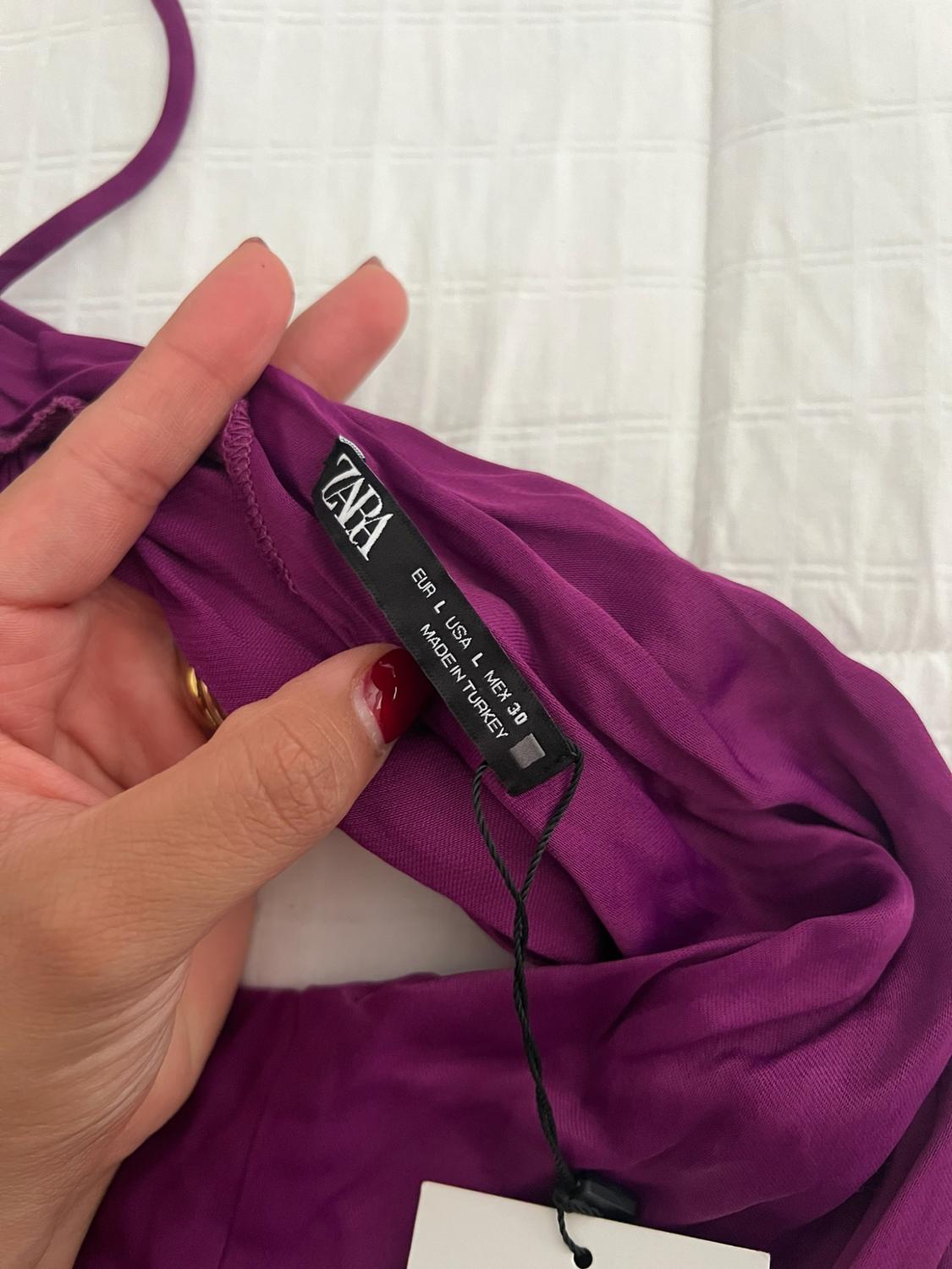 Hand holding a purple garment showing a label with the text ZARA EUR L USA L MEX 30 MADE IN TURKEY against a white background.