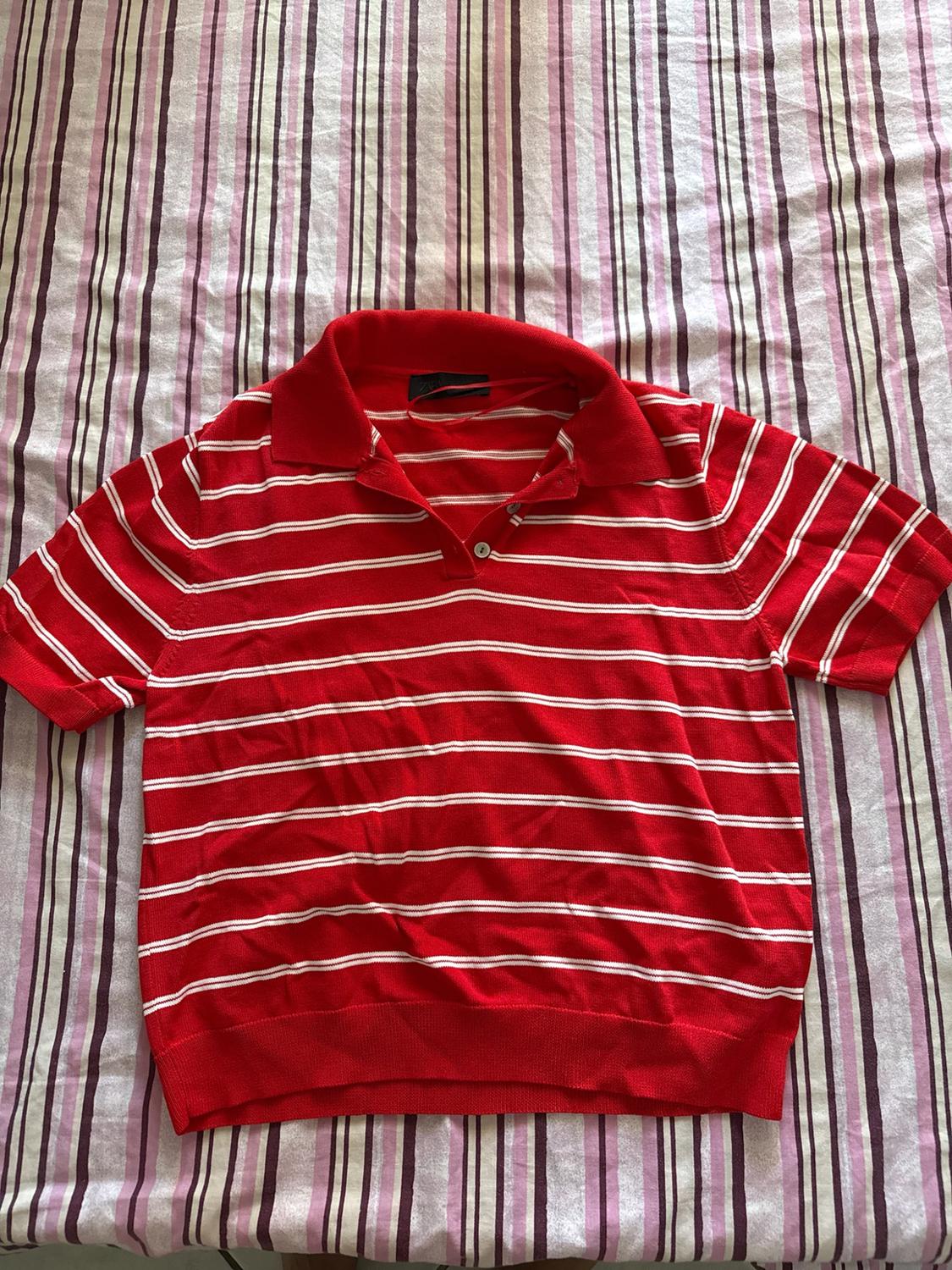 Short-sleeved red polo shirt with white horizontal stripes and a button-down collar, against a vertically striped fabric background.